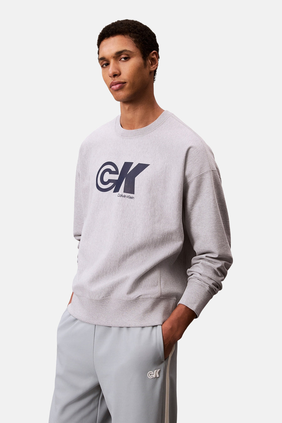 Sweat-shirt gris par Calvin Klein, avec un logo sur le devant et porté avec un pantalon de survêtement assorti.