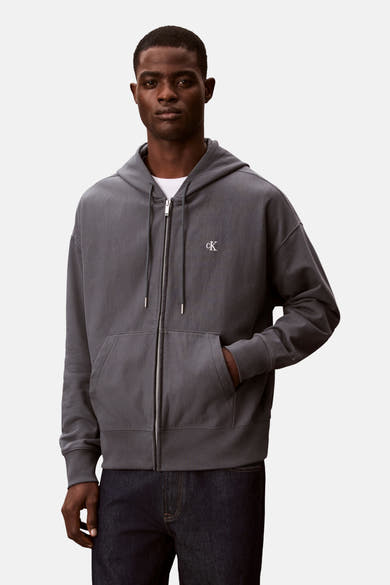 Blauwe Calvin Klein bodywarmer, met ritssluiting en capuchon, gedragen over een wit T-shirt en een donkere jeans.