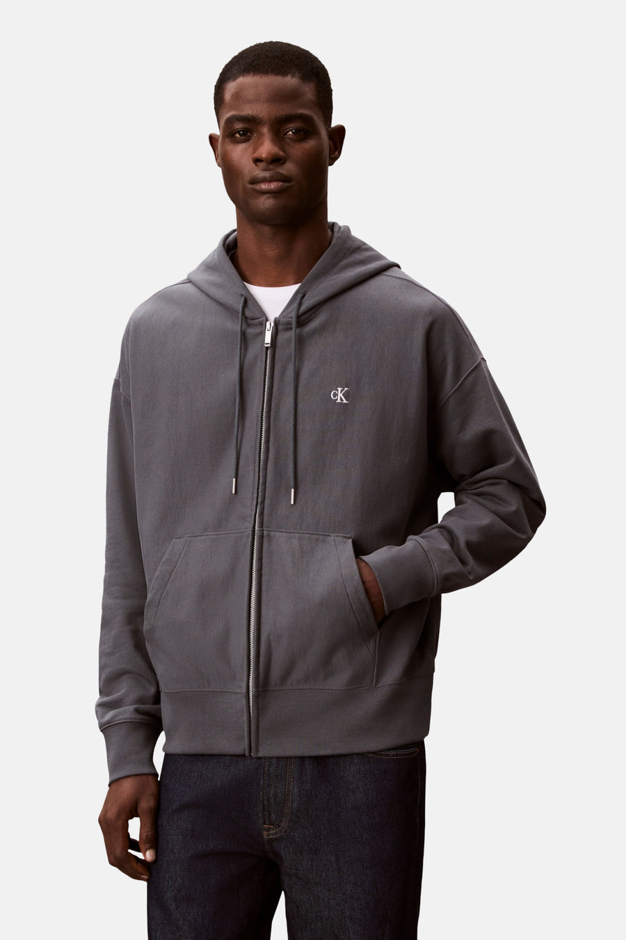 Blauwe Calvin Klein bodywarmer, met ritssluiting en capuchon, gedragen over een wit T-shirt en een donkere jeans.