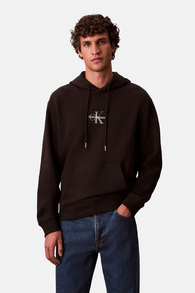 Zwarte Calvin Klein hoodie, gedragen met een blauwe spijkerbroek, op een effen achtergrond.