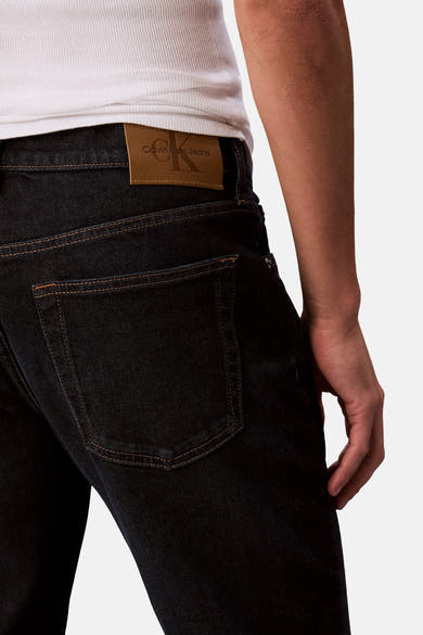 Jean fuselé bleu foncé de Calvin Klein, avec une étiquette en cuir apparente et une poche arrière sur un fond blanc.