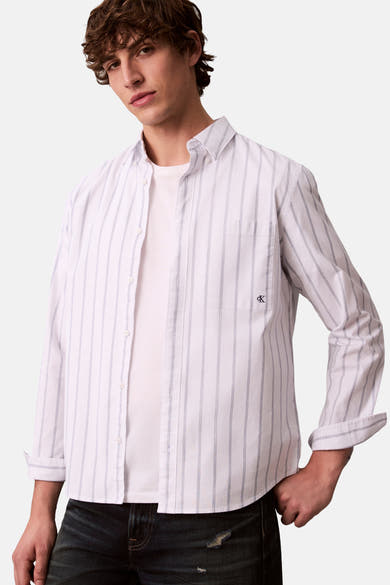 Chemise à manches longues blanche à rayures de Calvin Klein, portée ouverte sur un t-shirt uni, en jean foncé.
