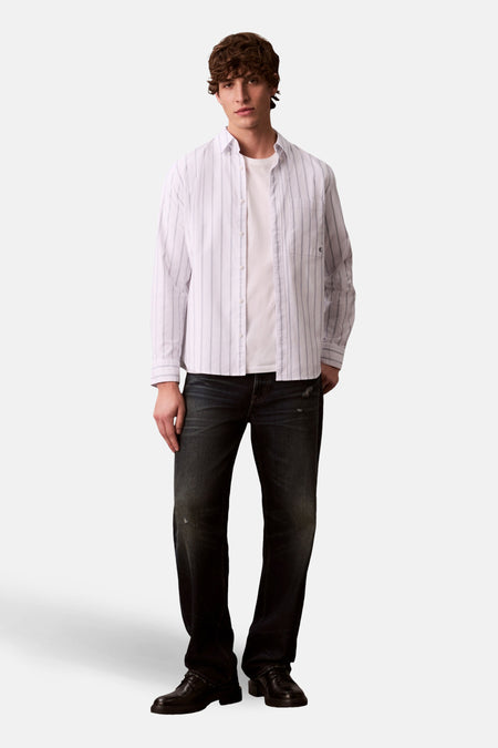 Witte lange mouw chemise met verticale strepen, van Calvin Klein, gedragen over een wit t-shirt en een donkere broek.