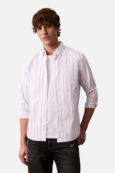 Chemise à manches longues blanche de Calvin Klein, rayures verticales bleu-gris, portée ouverte sur un t-shirt blanc et un jean foncé.
