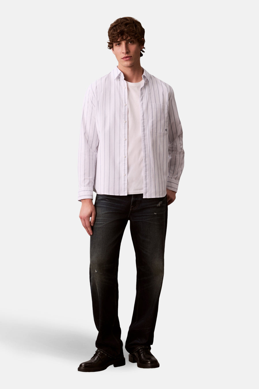 Chemise à manches longues blanche à rayures, de Calvin Klein, portée sur un t-shirt blanc avec un jean foncé et des chaussures noires.