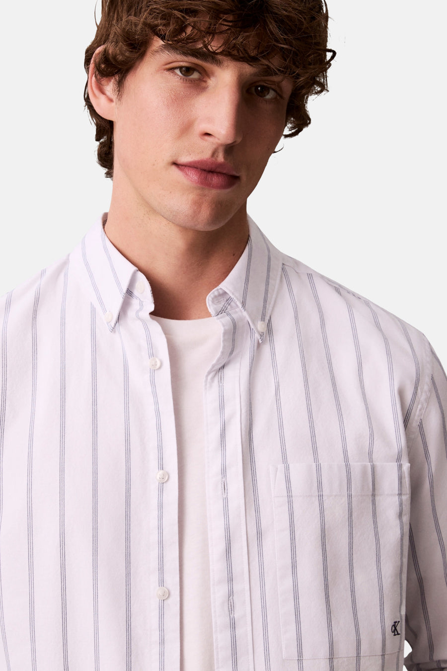 Chemise blanche à rayures verticales de Calvin Klein, portée sur un t-shirt blanc uni, avec logo sur la poche.