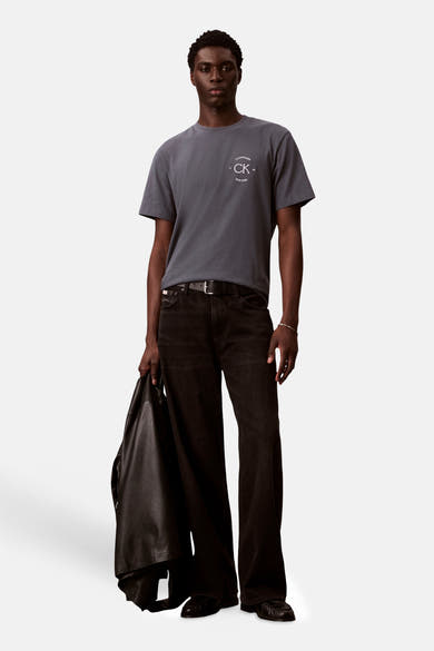 Grijs T-shirt met korte mouwen van Calvin Klein, met opdruk van het logo, gedragen met een zwarte broek en een jas.