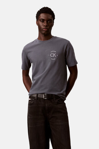 T-shirt gris à manches courtes de Calvin Klein, avec un logo décoratif sur la poitrine, porté avec un jean noir et une ceinture.