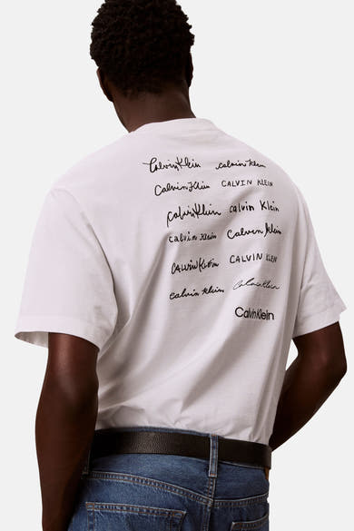 T-shirt blanc à manches courtes de Calvin Klein avec imprimé logo manuscrit au dos, porté avec un jean bleu et une ceinture noire.
