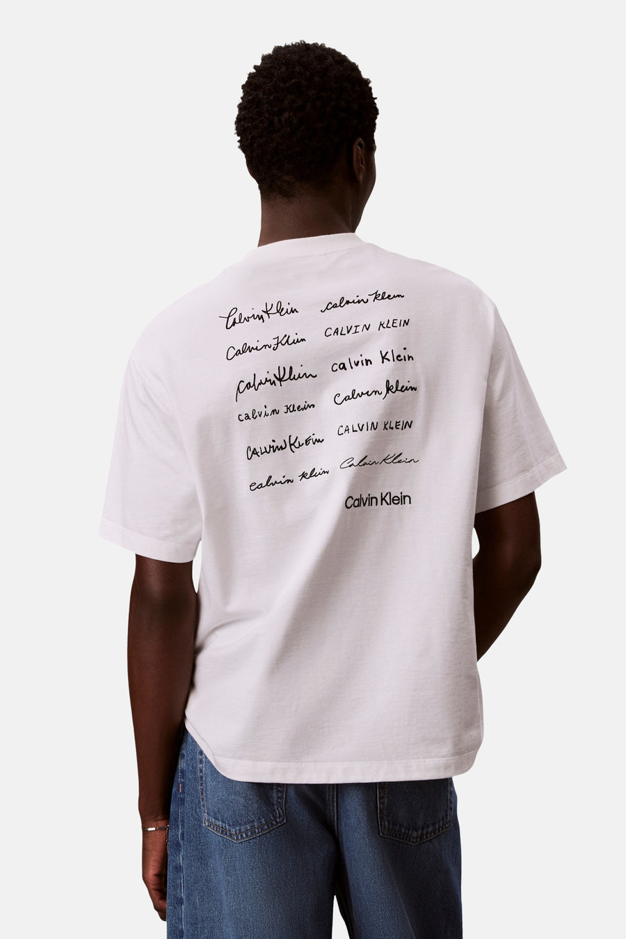 T-shirt blanc à manches courtes de Calvin Klein, orné d'un imprimé de logo multiple au dos, porté avec un jean bleu.
