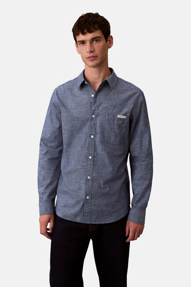 Chemise bleu à manches longues de Calvin Klein avec poche poitrine et texture subtile, portée avec un pantalon noir.
