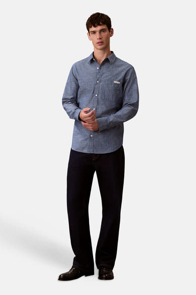 Chemise bleu à manches longues de Calvin Klein, avec une poche poitrine et portée avec un jean foncé et des chaussures noires.
