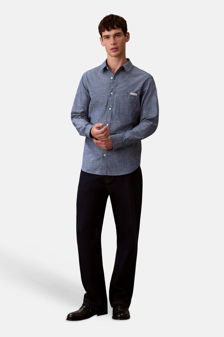 Chemise bleu à manches longues de Calvin Klein, avec une poche poitrine et portée avec un jean foncé et des chaussures noires.
