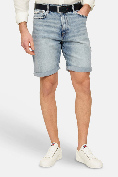 Short en jean bleu clair de Calvin Klein avec ourlet retroussé, porté par un mannequin avec une chemise et des baskets blanches.
