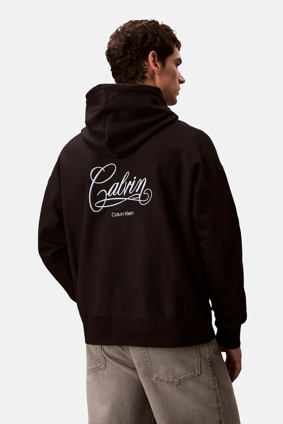 Sweat à capuche noir de Calvin Klein, avec logo cursif imprimé au dos, porté avec un jean gris.
