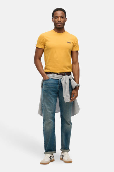 T-shirt à manches courtes ocre de SUPERDRY, porté avec un jean bleu et une chemise rayée nouée à la taille.