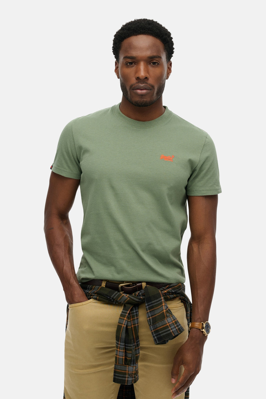 T-shirt kaki à manches courtes de SUPERDRY, porté avec un pantalon beige et une chemise à carreaux nouée à la taille.