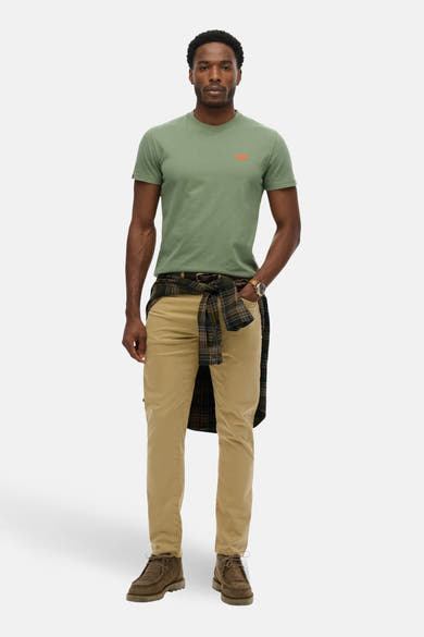 T-shirt kaki à manches courtes de SUPERDRY, porté avec une chemise à carreaux nouée, un pantalon beige et des bottines marron.