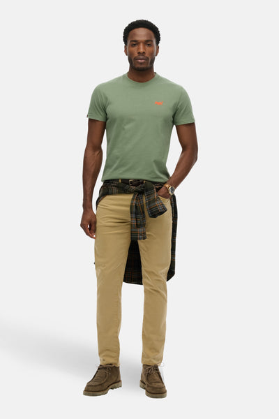 Kaki T-shirt met korte mouwen van SUPERDRY, gedragen met een geruit overhemd dat is vastgebonden, een beige broek en bruine enkellaarsjes.