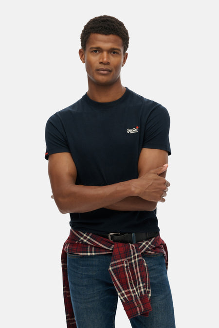 T-shirt bleu à manches courtes de SUPERDRY, porté avec une chemise à carreaux nouée à la taille et un jean.