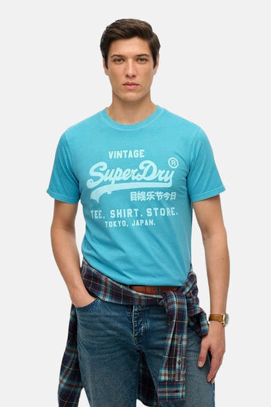 T-shirt bleu à manches courtes de SUPERDRY, avec un imprimé graphique, porté avec une chemise à carreaux et un jean.
