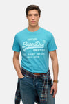 T-shirt bleu à manches courtes de SUPERDRY, avec un imprimé graphique, porté avec une chemise à carreaux et un jean.
