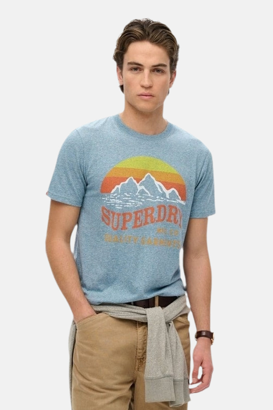 Blauw T-shirt met korte mouwen van SUPERDRY, met een berg- en zonmotief, gedragen met een beige broek en een geknoopte trui.