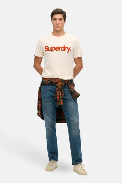 Beige T-shirt met korte mouwen van Superdry, gedragen met een geruit overhemd dat aan de taille is geknoopt en een spijkerbroek.