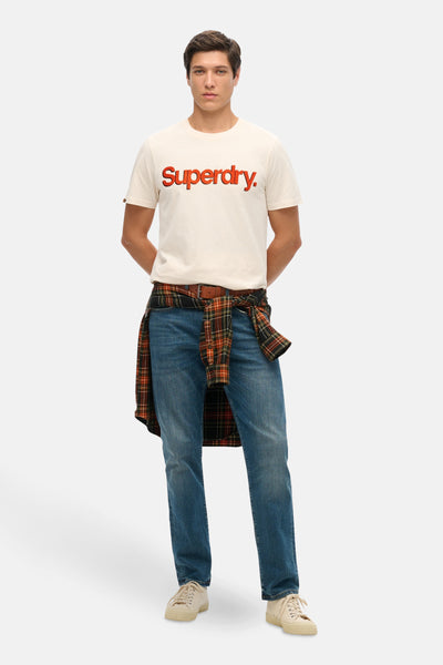 Beige T-shirt met korte mouwen van Superdry, gedragen met een geruit overhemd dat aan de taille is geknoopt en een spijkerbroek.