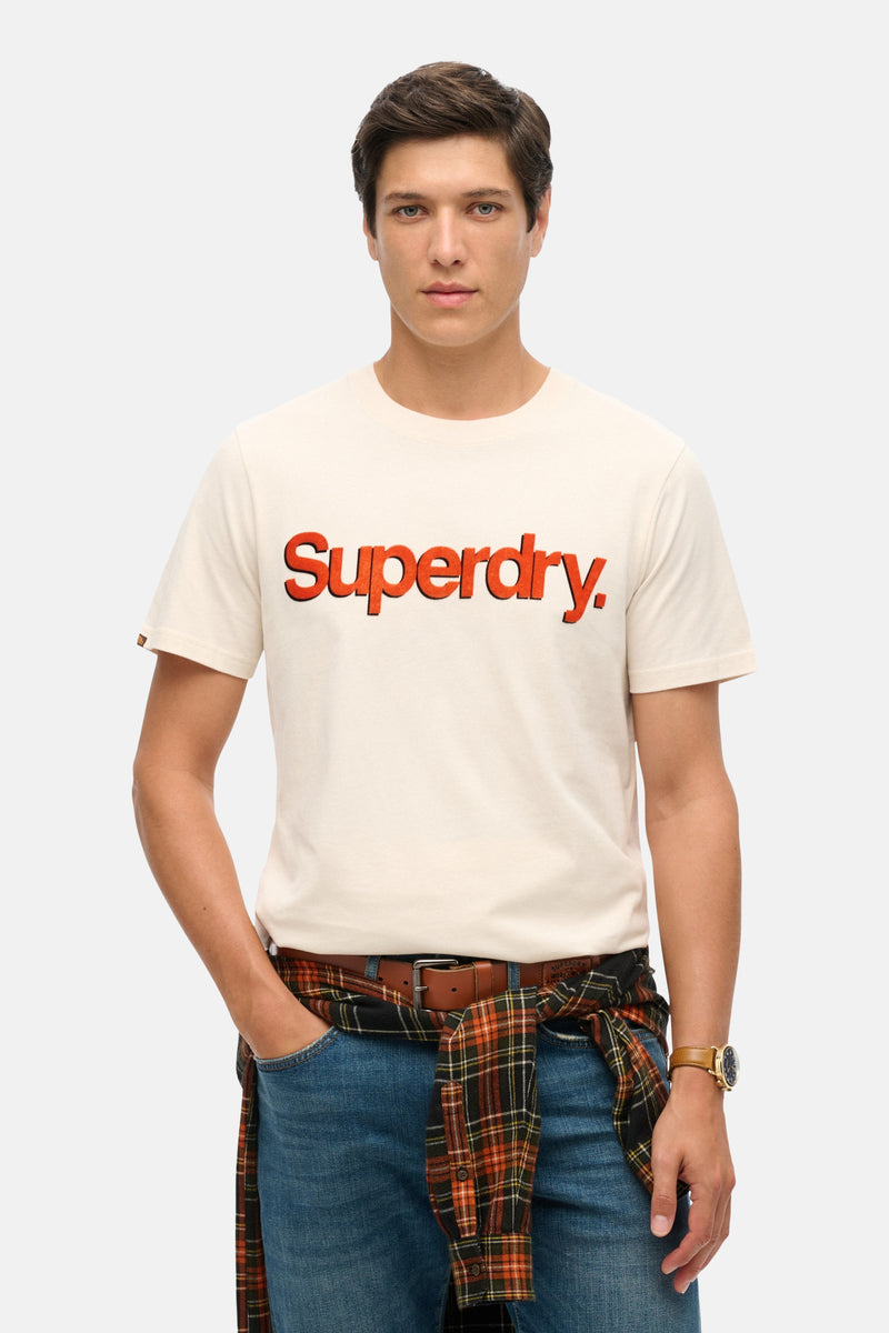 Beige T-shirt met korte mouwen van SUPERDRY, gedragen met een geruit overhemd en een spijkerbroek.