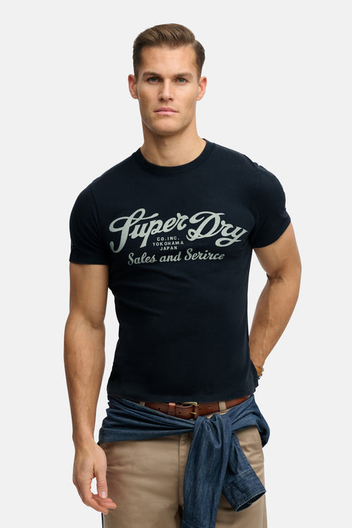 T-shirt bleu à manches courtes de SUPERDRY, avec inscription stylisée sur le devant, porté avec un pantalon beige et une chemise nouée.