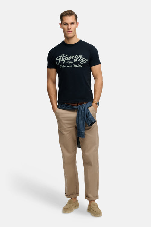 T-shirt bleu à manches courtes de SUPERDRY, avec un jean noué à la taille et un pantalon chino beige.