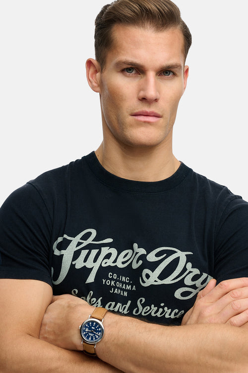 T-shirt bleu à manches courtes de SUPERDRY, porté par un homme aux bras croisés.
