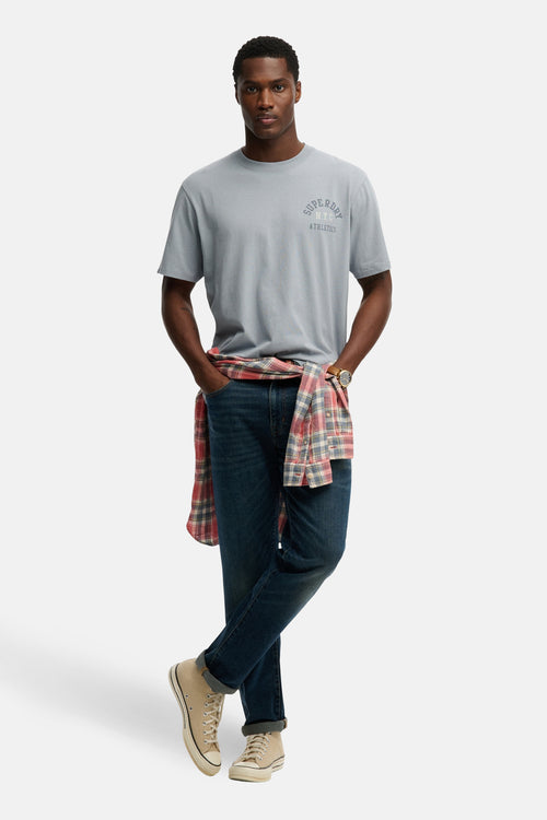 T-shirt bleu à manches courtes de Superdry, avec un imprimé sur la poitrine, porté avec une chemise à carreaux et un jean.
