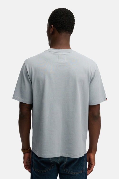 T-shirt bleu à manches courtes de SUPERDRY, avec un ourlet rouge sur la manche et porté avec un bracelet marron et un jean bleu.