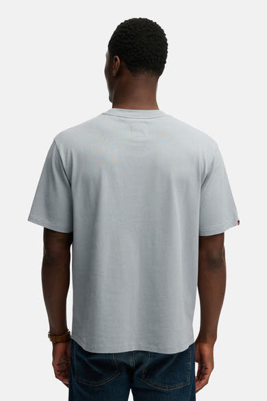 Blauw T-shirt met korte mouwen van SUPERDRY, met een rode zoom op de mouw en gedragen met een bruine armband en een blauwe spijkerbroek.