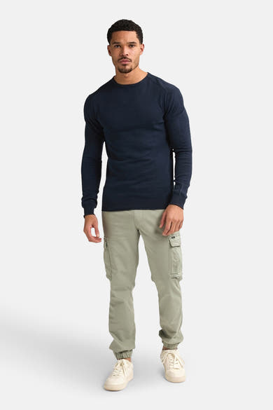 Pull à col rond bleu de Petrol Industries, porté avec un pantalon cargo vert olive et des baskets couleur crème.