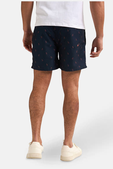 Short de bain bleu foncé de Petrol Industries®, à motif de palmiers rouges, porté avec un t-shirt blanc et des chaussures blanches.