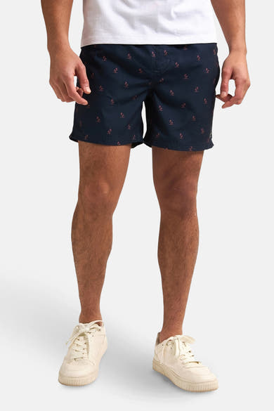 Short de bain bleu foncé de Petrol Industries®, avec motif de palmiers stylisés et porté avec des chaussures blanches.
