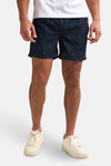 Short de bain bleu foncé de Petrol Industries®, avec motif de palmiers stylisés et porté avec des chaussures blanches.
