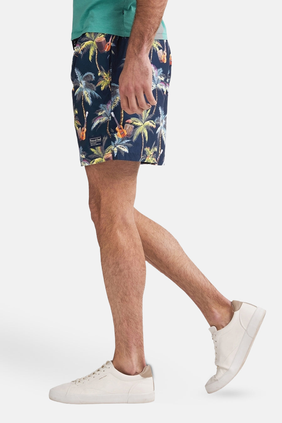 Blauwe zwembroek van Petrol Industries, bedrukt met palmbomen en gitaren, gedragen met een turquoise T-shirt en witte sneakers.