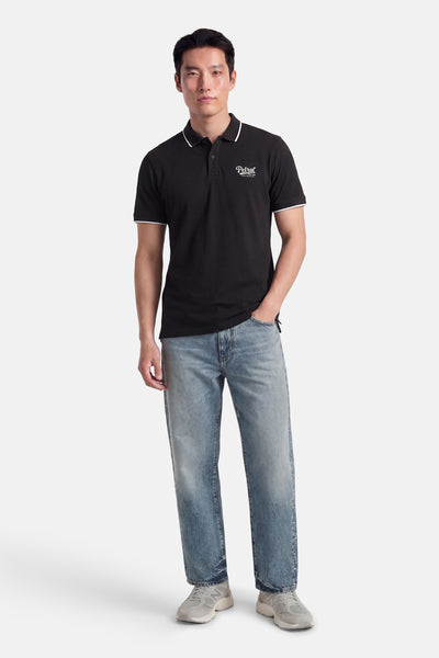 Polo noir à manches courtes de Petrol Industries, avec un col et des ourlets blancs, porté avec un jean délavé et des baskets grises.