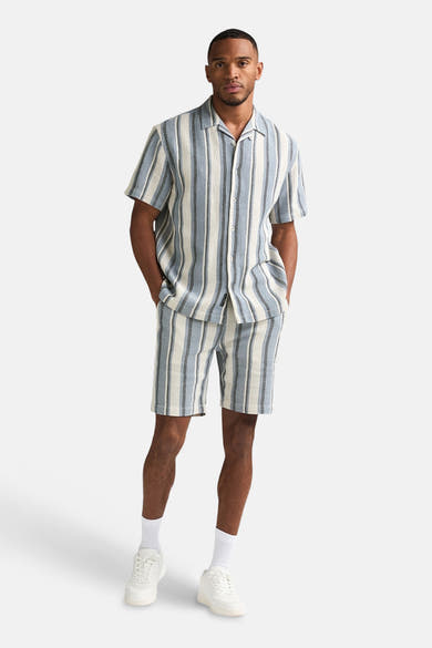 Korte blauwe short van Petrol Industries®, met verticale strepen, gedragen met een bijpassend overhemd en witte sneakers.