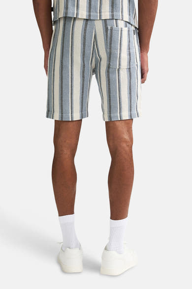 Korte blauwe gestreepte broek van Petrol Industries, achterkant, met witte en zwarte verticale strepen, gedragen met witte sokken en witte sneakers.