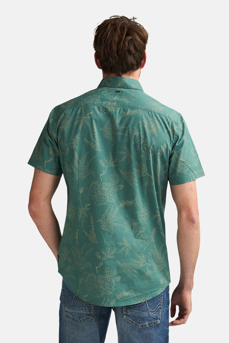 Chemise à manches courtes verte de Petrol Industries, avec un motif tropical imprimé, portée avec un jean bleu.
