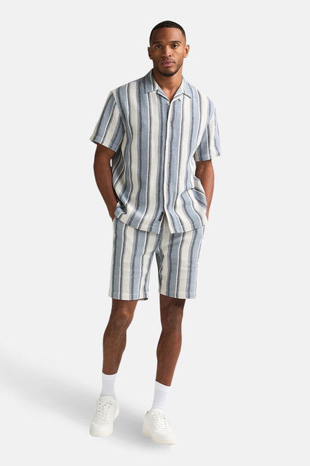 Chemise à manches courtes bleue rayée de Petrol Industries, portée avec un short assorti, des chaussettes blanches et des baskets blanches.