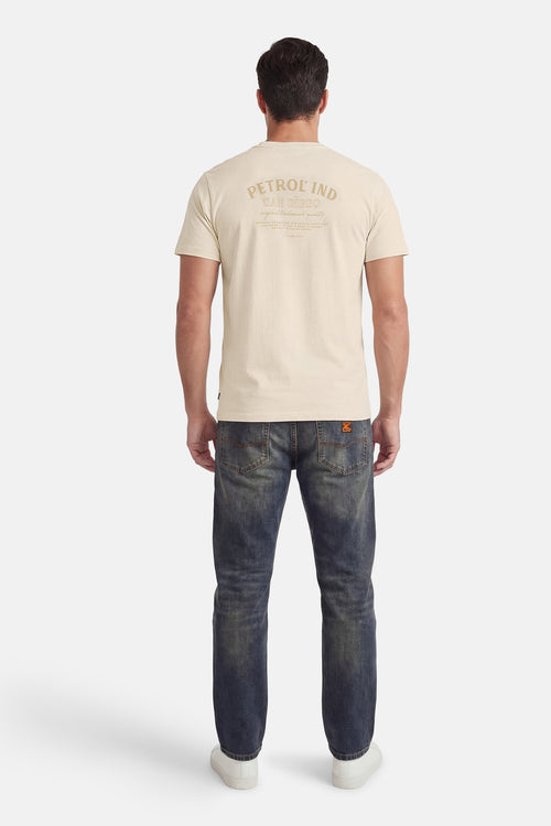 T-shirt écru à manches courtes de Petrol Industries, avec un imprimé texte décoratif au dos, porté avec un jean bleu et des baskets blanches.
