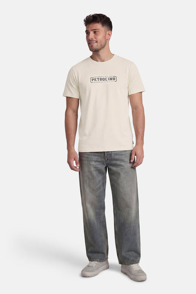 T-shirt à manches courtes écru de Petrol Industries, avec un logo graphique sur le devant, porté avec un jean ample et des chaussures de sport.