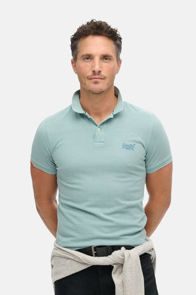 Polo bleu à manches courtes de SUPERDRY, porté avec un pull gris noué autour de la taille.