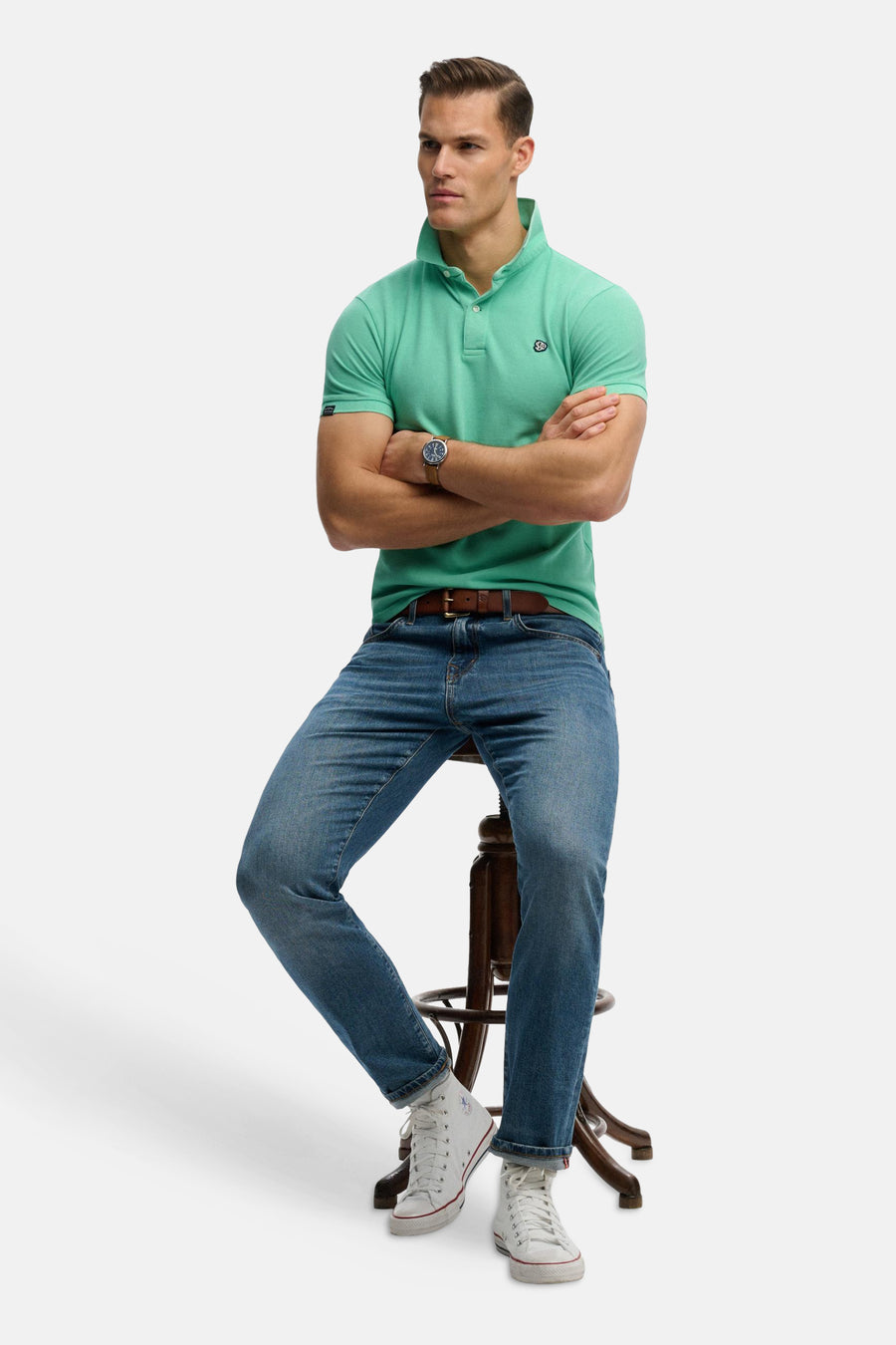 Groen SUPERDRY poloshirt, gedragen met een blauwe jeans, een bruine riem en witte sneakers.